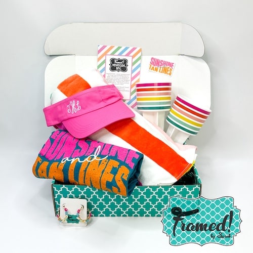 Framed Monogram Box open and displaying the July contents. Items include, striped towel, colorful striped sand cup holders, hot pink visor with white monogram, Teal "sunshine and Tan Lines" t-shirt, beaded daisy earrings