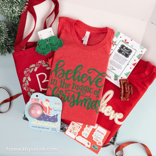 Open Monogram Box™ displaying the items inside. Contents include: red Believe in the Magic of Christmas t-shirt, red Believe sweatshirt, monogrammed red wreath tote, holiday bath bomb, green fringe earrings, red hair claw, and holiday gift tag sticker sheet